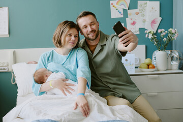 Obraz premium Happy couple in hospital room taking selfie while holding newborn baby. Background displaying posters, plants, and personal items, enhancing the intimate and joyful family moment