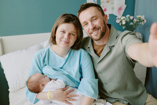 Portrait of family holding newborn baby in hospital room. Parents smiling while mother wearing hospital gown and holding swaddled infant