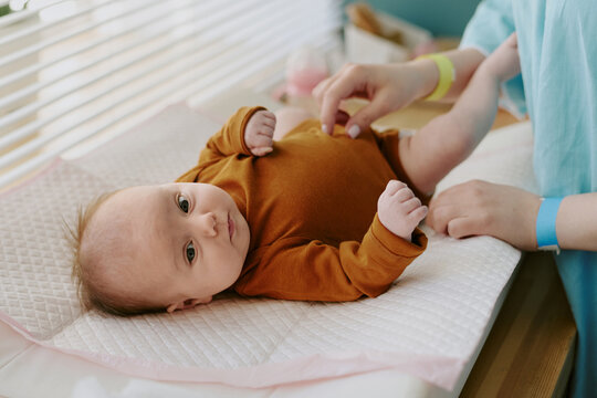 Portrait of baby lying on changing table while caregivers hands adjusting babys clothing. Baby glancing up with curiosity in face, showcasing calm and peaceful expression