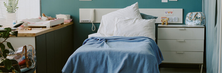 Cozy hospital room featuring neatly made bed with blue blanket, bedside table with medical supplies, sunlight from window creating warm atmosphere
