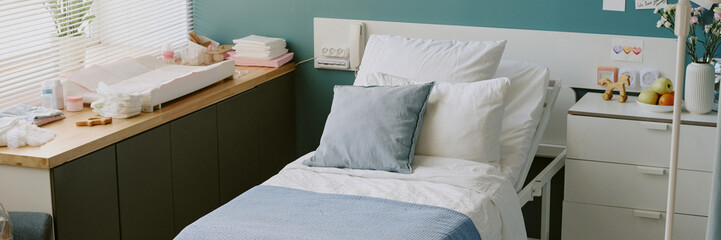 Hospital room featuring a bed with clean white linens and light blue blanket, accompanied by bedside table, window with blinds ushering sunlight and variety of medical equipment