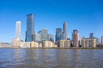 Fototapeta premium Panoramic view of Canary Wharf, showcasing modern skyscrapers and residential buildings. The clear blue sky enhances the vibrant scene along the River Thames in London.