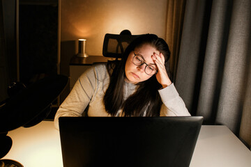 Woman working on computer late evening at home office. Concentrated female freelancer trying to...