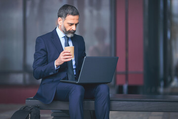 Senior businessman outdoors having coffee and using laptop