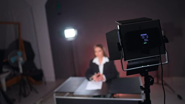 Lighting equipment back view at studio. Woman in black jacket and white shirt sits at blurred backdrop.