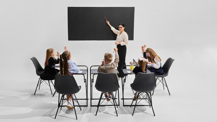 Teacher pointing at blackboard as all children actively raise hands to answer question. Concept of interactive learning, school engagement, teamwork, back to school, participation, school enthusiasm.