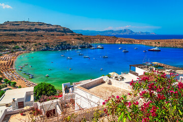 Lindos, Rhodes, Greece:  Scenic view of idyllic Lindos Bay with Clear Blue Waters and White Sailboats, Nestled Between Dramatic Cliffs and a Charming Coastal Village