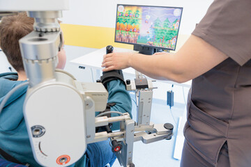 Child receiving robotic arm therapy while interacting with a game on screen in a pediatric rehab clinic