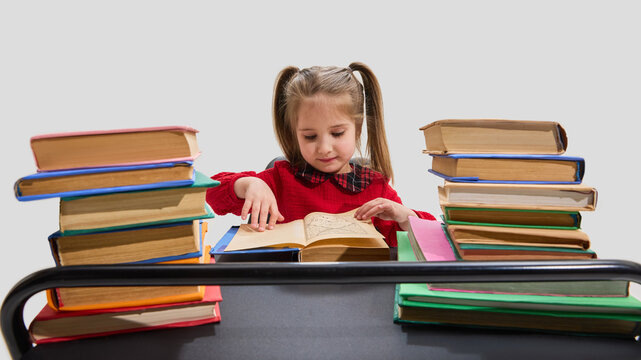 Girl in red sweater surrounded by tall stacks of books focuses on reading open textbook on desk against white background. Concept of education, library study, knowledge, early learning, and curiosity.
