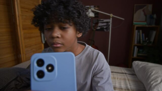Adorable African American child sitting on bed and looking at smartphone screen while suddenly seeing indecent content in the internet, turning his head back and smiling