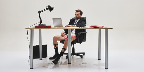 Full length photo of young man in suit jacket and underwear working from home at desk with laptop...