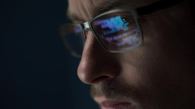 Close up of serious software engineer wearing glasses looks at his screen, with code reflecting on his eyeglasses. Coder is focused on programming, data analysis, or cybersecurity