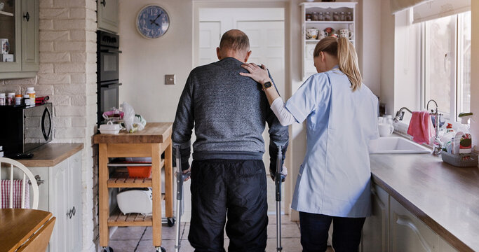 Nurse support senior man in crutches in home kitchen, medical rehabilitation and recovery. Caregiver back, person with a disability walk and help, health and physiotherapy, healing injury or accident