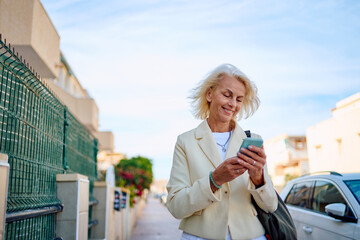 Older woman smiles while using smartphone on sunny street in residential area
