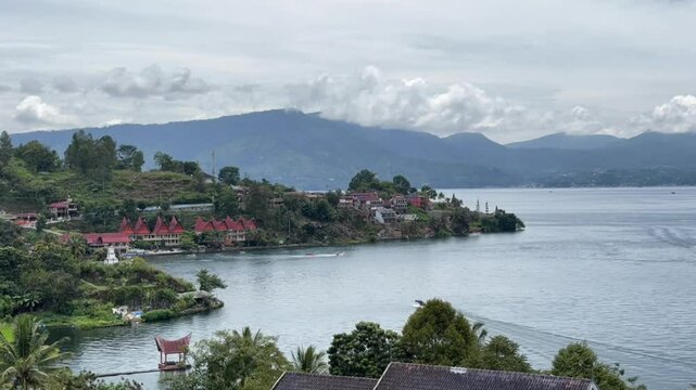 Timelapse footage of Lake Toba and TukTuk village in North Sumatra, Indonesia, showing calm waters, traditional Batak architecture, and surrounding lush mountains under a cloudy sky.