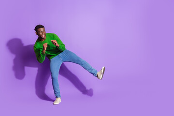 Young man in bright green sweatshirt with casual jeans dancing energetically on a purple background expressing joy and style