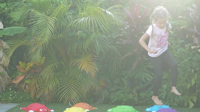 Two happy girls running and playing outside on artificial grass with colorful turtle stepping stones, tropical plants background, barefoot childhood activity, outdoor fun and movement, candid moment