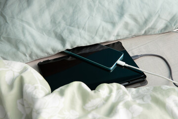 Smartphone and tablet computer are charging in bed under a pillow and blanket. Concept of overheat...