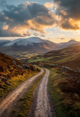 a winding dirt road leads towards snow-capped mountains under a dramatic and golden sunset sky in the irish countryside.