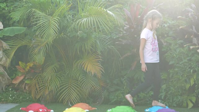 Little girl running and playing outside on artificial grass with colorful turtle stepping stones, tropical plants background, barefoot childhood activity, outdoor fun and movement, candid moment