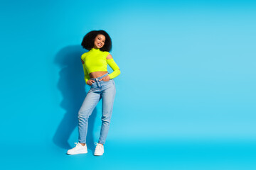 Smiling young woman in neon green top and jeans posing confidently against a vibrant blue...