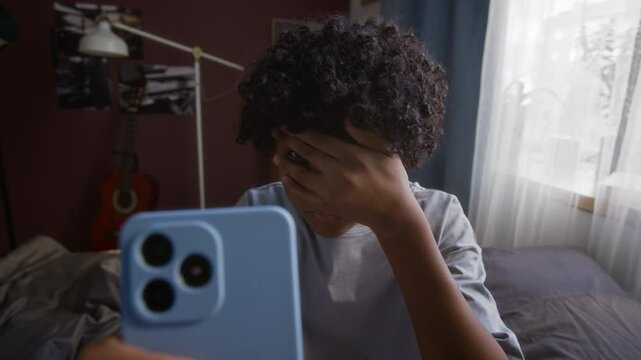 Shocked schoolboy covering his face by hand and looking through fingers at smartphone screen while accidentally saw indecent online advert or mature content