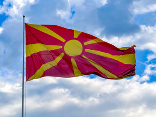 The vibrant flag of North Macedonia proudly flutters on a flagpole under a cloudy blue sky. The red and yellow colors stand out, symbolizing national pride