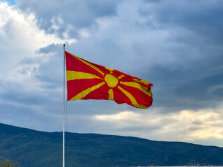 The Macedonian flag flies prominently against a backdrop of dark clouds and distant mountains, creating a striking visual contrast during the late afternoon. This captures a moment of national pride