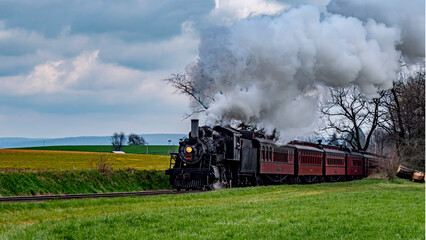 A steam locomotive chugs along the railway, releasing white smoke while passing lush green fields. The clouds overhead hint at a spring afternoon atmosphere.