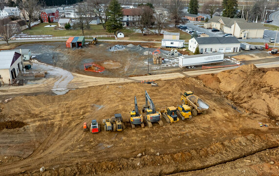 Heavy machinery including excavators and dump trucks is actively working at a construction site in a rural area. Workers are overseeing the excavation process in daylight.