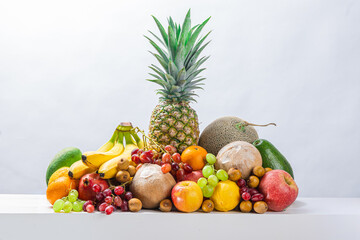 Colorful tropical fruits including pineapple, banana, melon, coconut, grapes, apple, orange, and mango arranged neatly on white surface with bright background