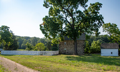 A rustic stone house stands amidst a verdant landscape, featuring a large tree nearby. A white fence frames the property, with a dirt path leading towards the house under clear blue skies.