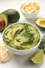 Tasty avocado dip in bowl, dill, lime and puffed rice cakes on white wooden table, closeup