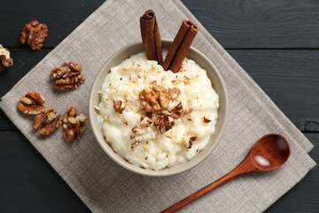 Delicious rice pudding with walnuts, cinnamon and spoon on black wooden table, flat lay