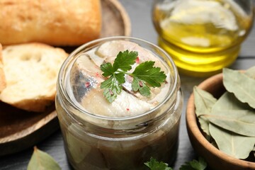 Pieces of delicious herring with spices in jar and bread on black table, closeup