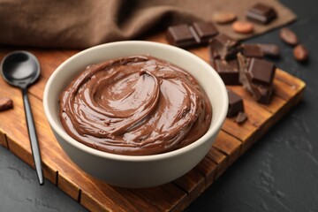 Tasty pudding in bowl, pieces of chocolate and cocoa beans on black table, closeup