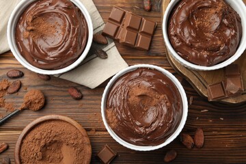Tasty pudding in bowls, pieces of chocolate and cocoa beans on wooden table, flat lay