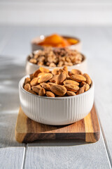 Three bowls of healthy snacks including dried apricots, walnuts, and almonds are arranged on a wooden board, offering a nutritious and delicious treat