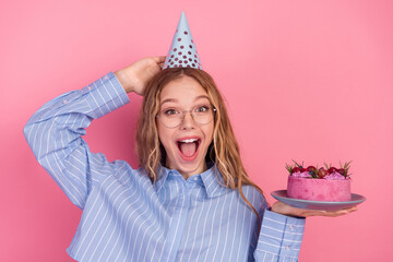 Excited young woman with party hat holding a cake on a pink background, celebrating joyfully with a vibrant expression