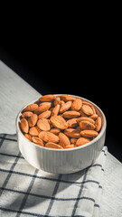 White bowl brimming with almonds resting on a checkered cloth atop a wooden table, creating a cozy, inviting atmosphere against a dark background. Perfect for highlighting healthy snacking