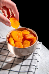 Hand is picking a dried apricot from a ceramic bowl full of dried apricots, placed on a checkered cloth on a white wooden table, with a black background