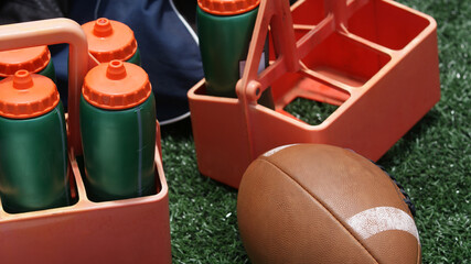 Football and water bottle on sideline symbolize game preparation, hydration, and sports training,...