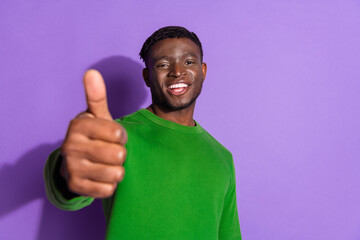 Smiling young man in a vivid green pullover demonstrating a positive thumbs up gesture against a vibrant purple background