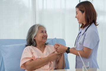 Smiling caring Asian doctor supporting holding hand of olde senior female patient at clinic or...