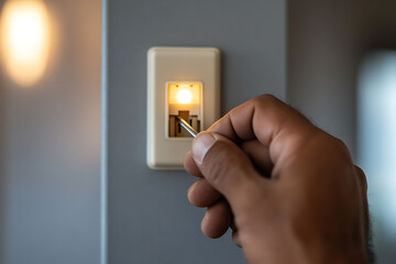 Dimmer switch adjustment: Man's hand uses a tool to control the light intensity of a slider-style dimmer switch on a gray wall. Interior lighting.