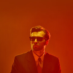 Man in sunglasses with confident chin-up pose against deep orange glow expressing cold detachment. Concept of ego, independence, masculinity, calm in chaos, emotion, mental state.