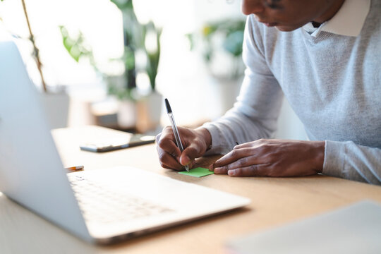 A young Arab man writes on a sticky note at his desk. He is working on his laptop in a bright office with plants. He is wearing a gray sweater.
