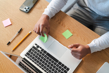 An Arab young man is working at his desk. He is putting a sticky note on his laptop as a reminder. The desk has a phone, pen, and pencil.
