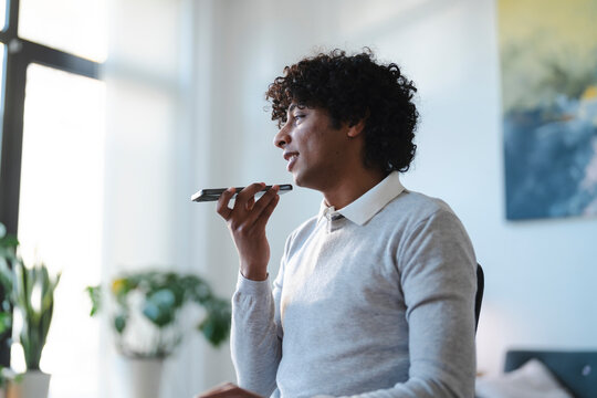 A young Arab man uses voice-to-text on his phone. He is sitting and holding the phone close to his mouth. He is wearing a gray sweater and a white collared shirt.