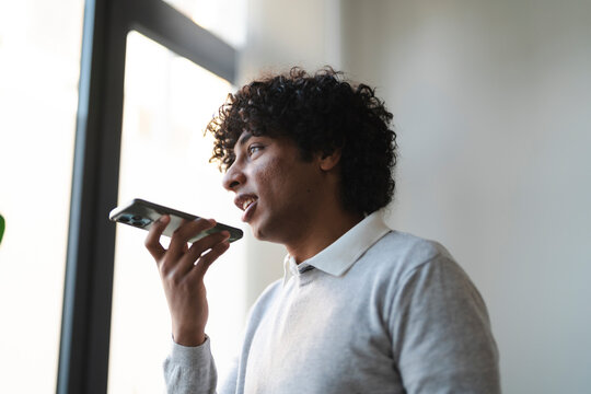 An Arab young man is using the voice command on his smartphone. He is standing near a window, wearing a gray sweater.